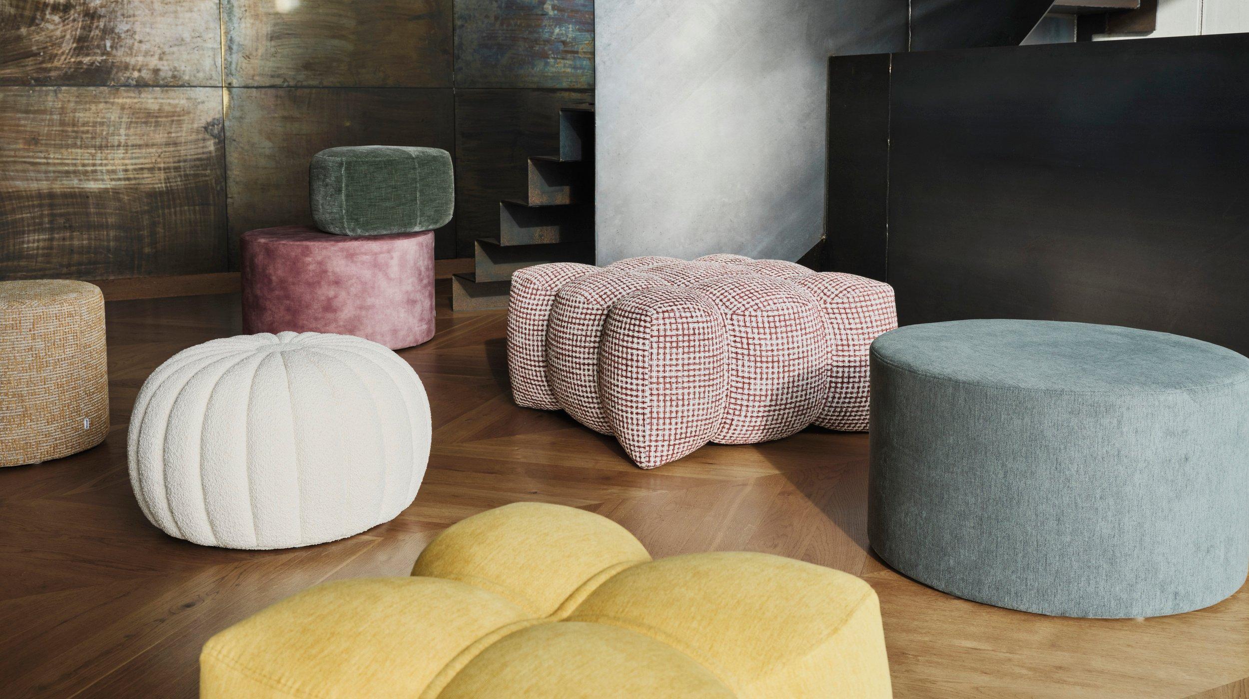 木地板上摆放着各种形状、颜色和纹理的脚凳组合，旁边是现代感楼梯和深色木饰面墙。